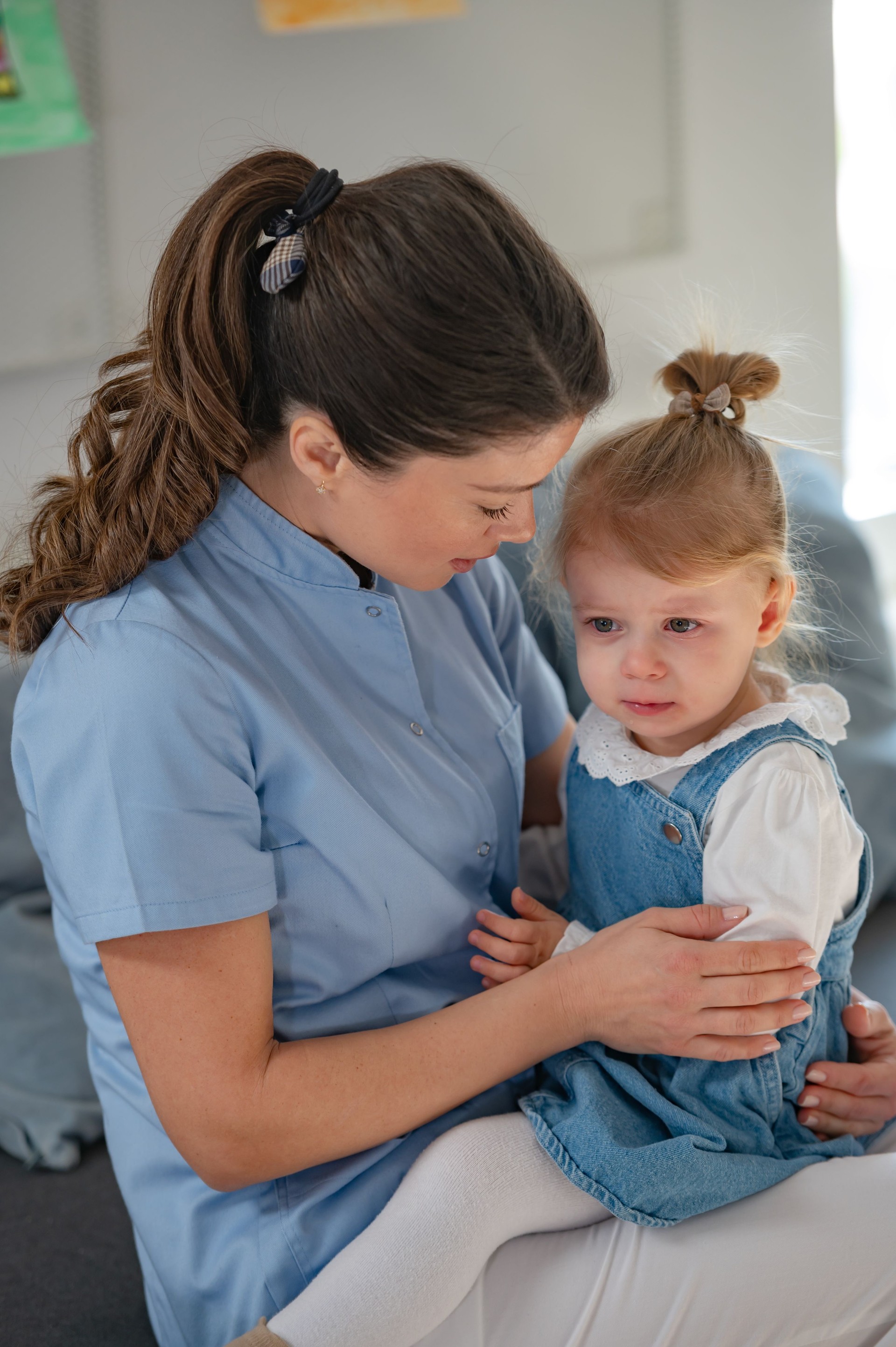 A caregiver in a blue uniform gently holds a sick young girl while comforting her on a couch, showing warmth and care in a classroom or daycare setting